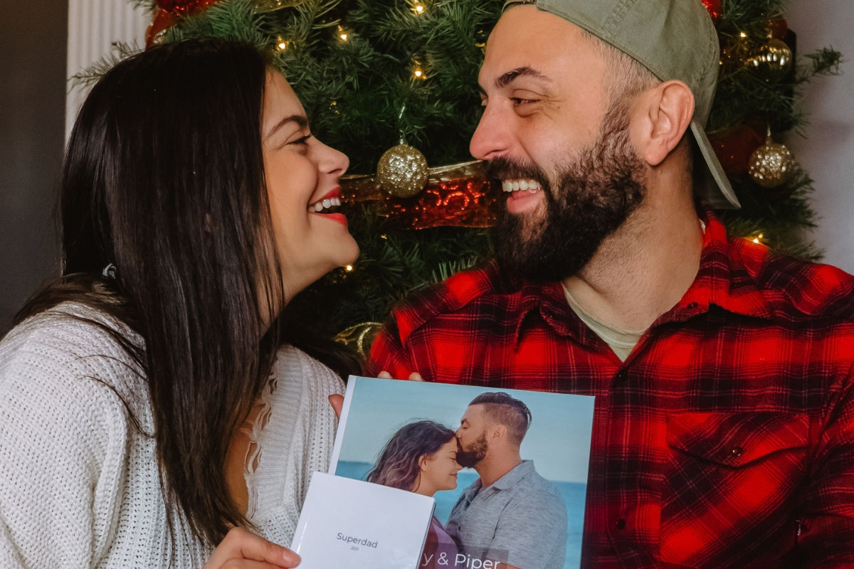 A laughing couple in front of a Christmas tree, holding a hardcover photo book with their own photo on the cover