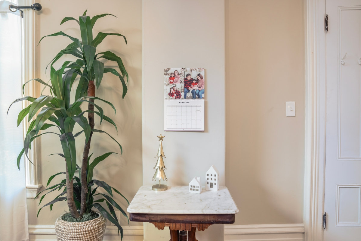 A wall calendar open to December, with a collage of Christmas family photos above the month grid, hanging over a marble side table with a gold Christmas tree and two small white houses