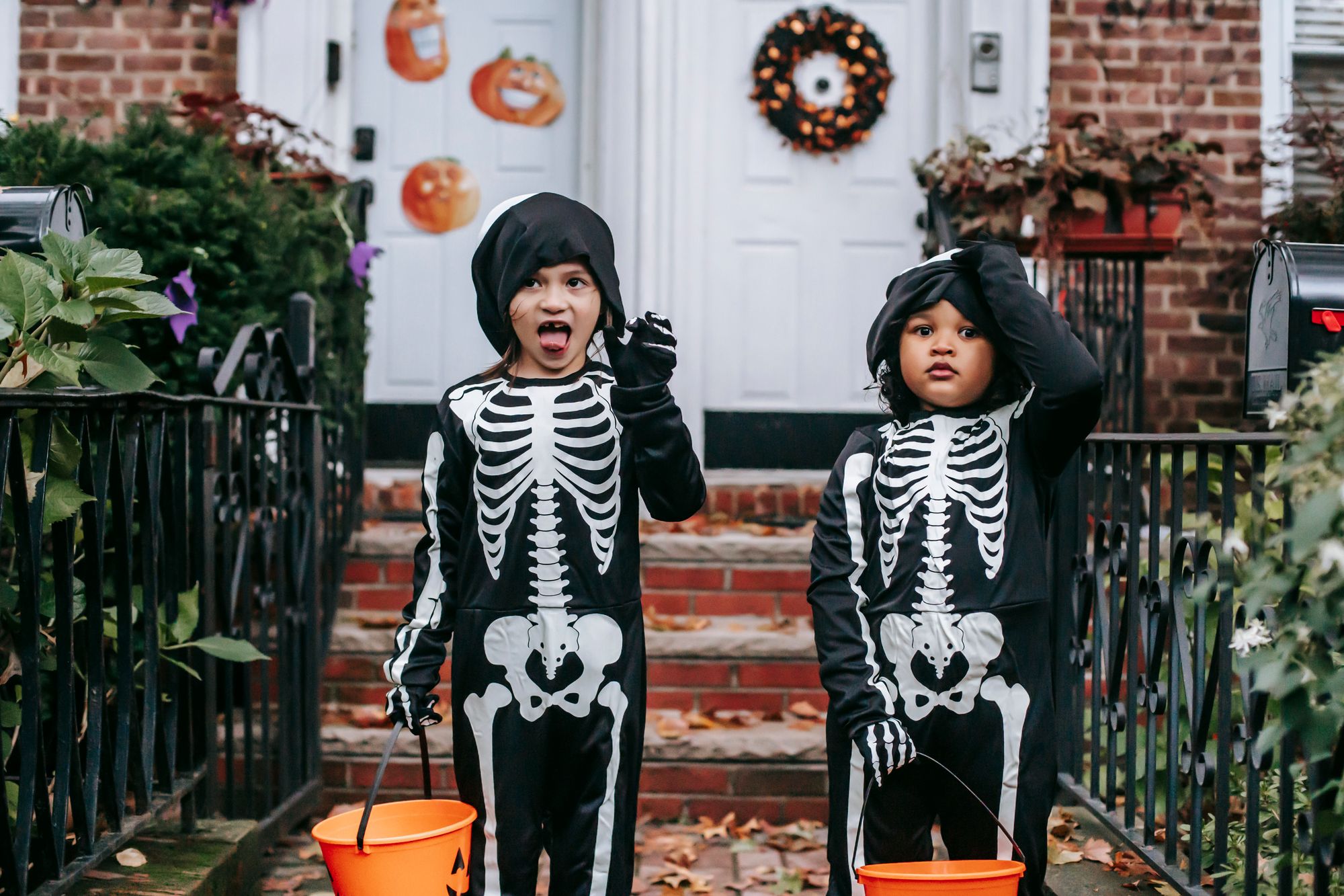 Two young children in matching skeleton costumes holding orange jack-o'-lantern buckets on the front path of a brick house decorated for Halloween