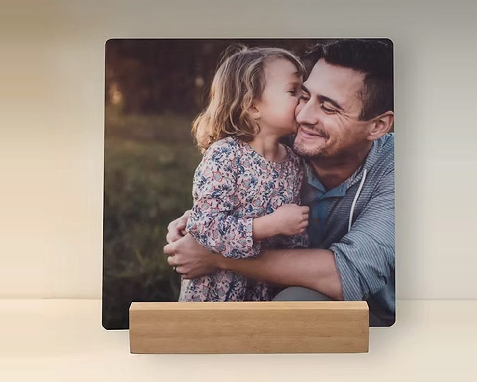 A metal desk print of a little girl kissing her father on the cheek, on its wooden stand