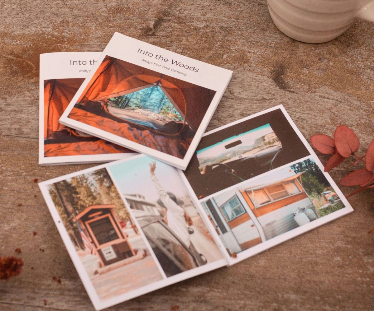 Two closed and one open mini photo book of a camping trip, on a wooden table beside a mug of coffee
