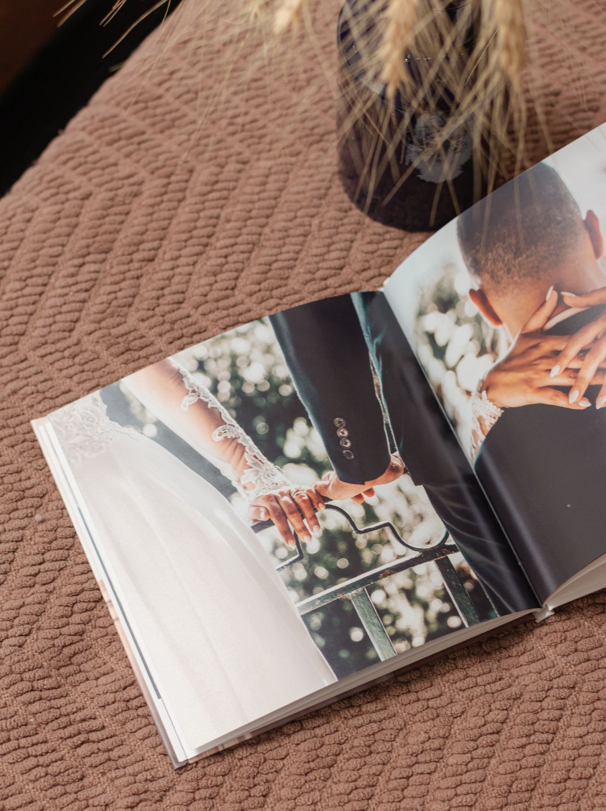 An open wedding photo book on a textured blanket, with a couple's joined hands on a railing