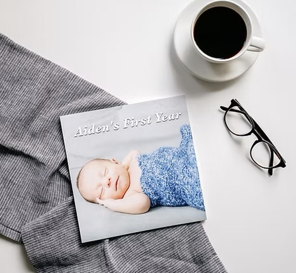 A softcover photo book titled Aiden’s First Year on a grey throw, beside a cup of coffee and reading glasses
