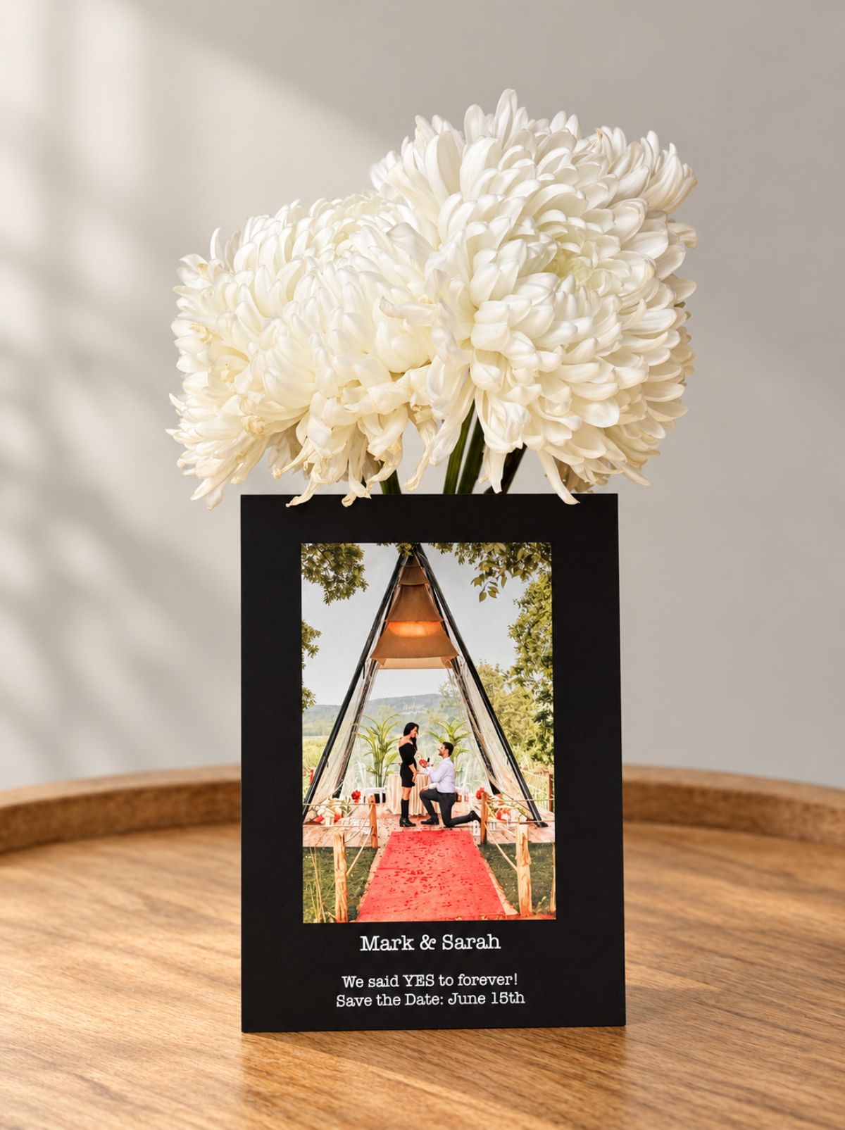 A black save-the-date photo card with a marriage proposal photo, standing in front of white chrysanthemums
