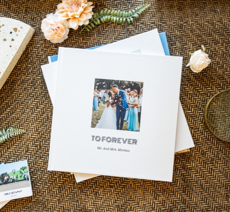 A 12x12 white hardcover wedding photo book titled To Forever, on a woven table with flowers