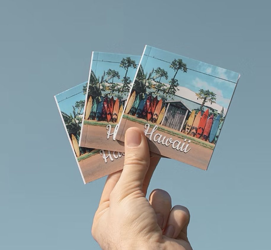 A hand holding up three mini photo books of a trip to Hawaii against a blue background