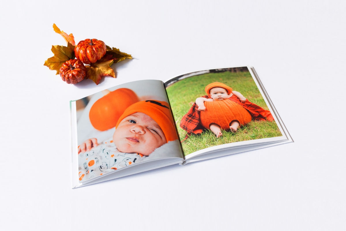 An open SimplePrints photo book showing two pages of baby photos in pumpkin costumes, next to small decorative pumpkins and autumn leaves