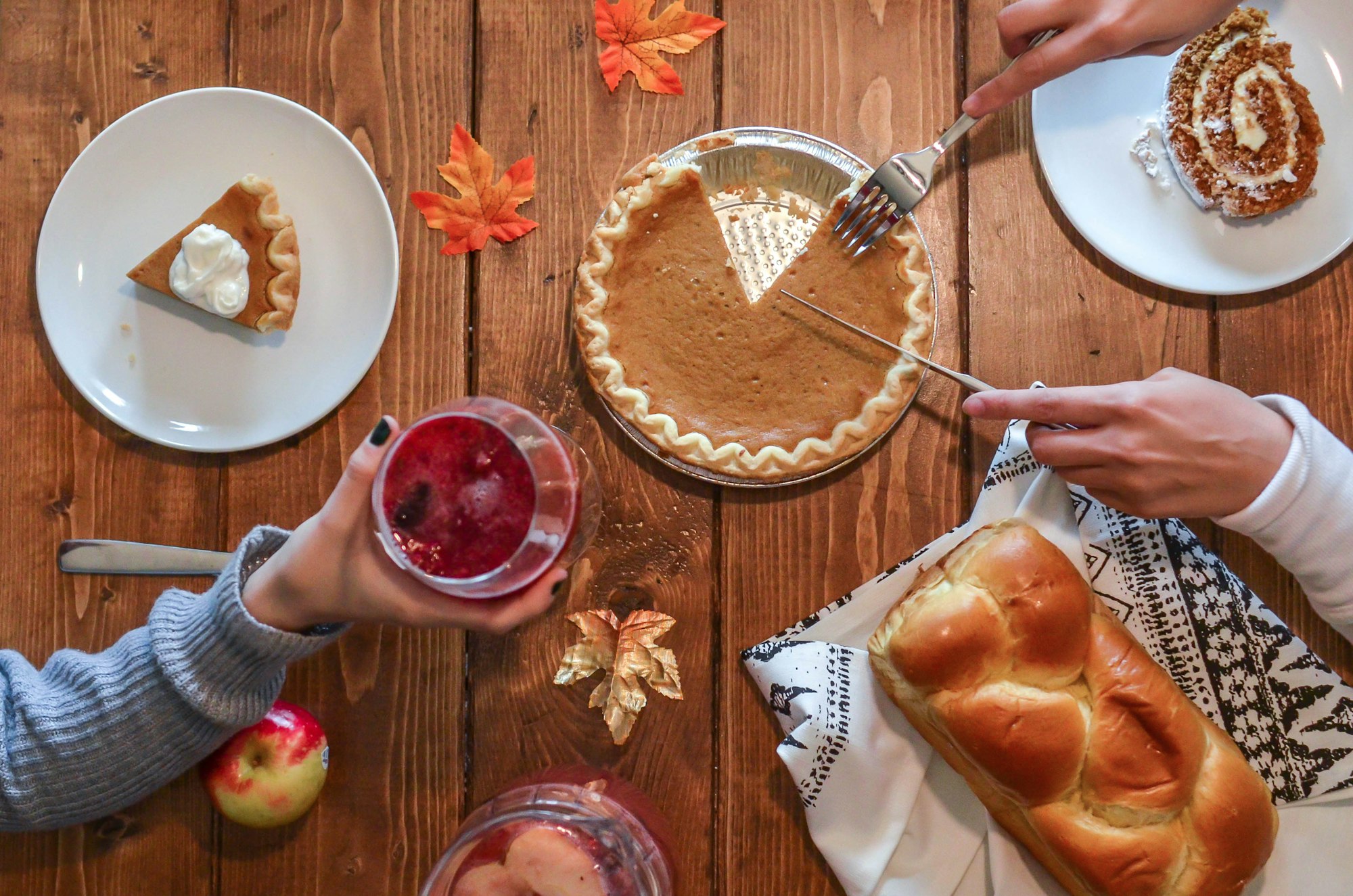 Friends sharing pumpkin pie, a pumpkin roll and braided bread on a wooden table scattered with autumn leaves