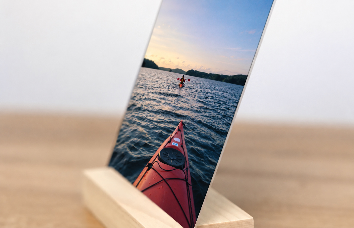 Side view of a travel desk print of a red kayak on a lake, resting in its solid wood display stand